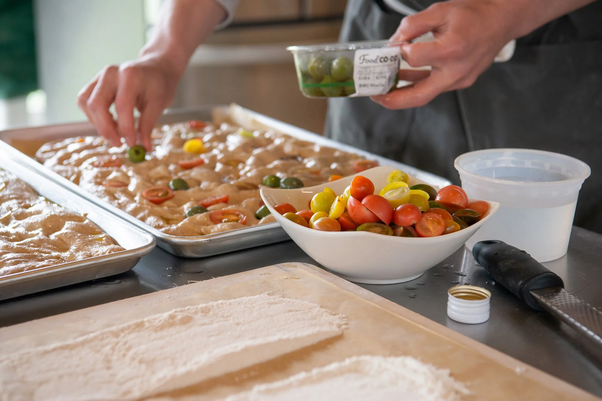 A person preparing cherry tomatoes and chicken for a dish in a kitchen. There are trays, a bowl of cut cherry tomatoes, a container of green grapes, and a piping bag on the counter.