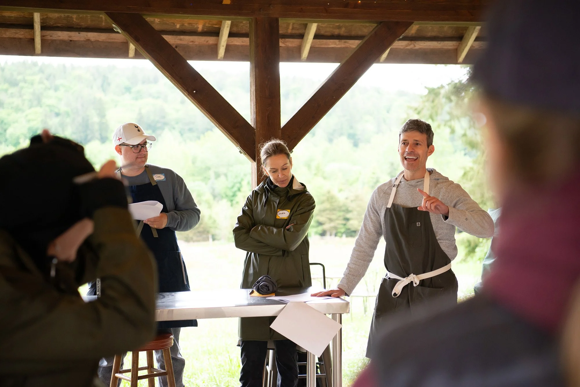 People gathered outdoors under a wooden pavilion, engaged in a discussion or presentation, with a man in a gray hoodie and apron speaking, while others listen and take notes.