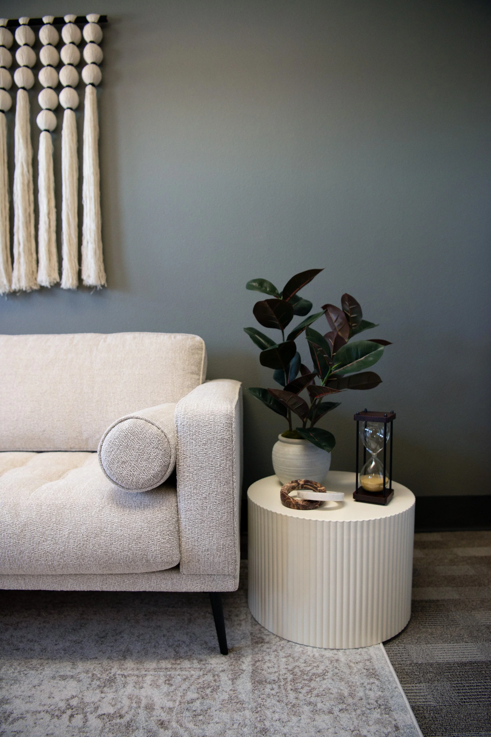 Living room with beige sofa, textured round pillow, white ribbed side table, potted houseplant, hourglass, and decorative tray.