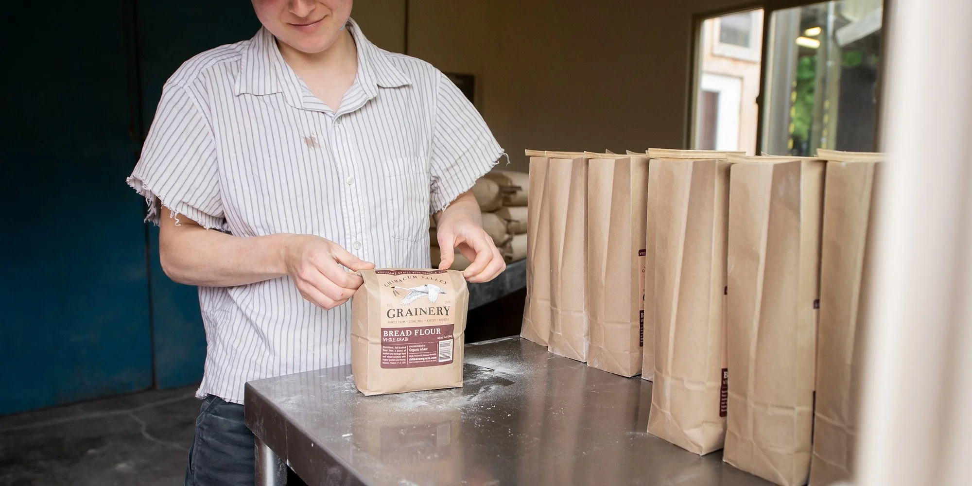 Person handling a paper bag labeled 'bread flour' in a room with a table and several other paper bags.