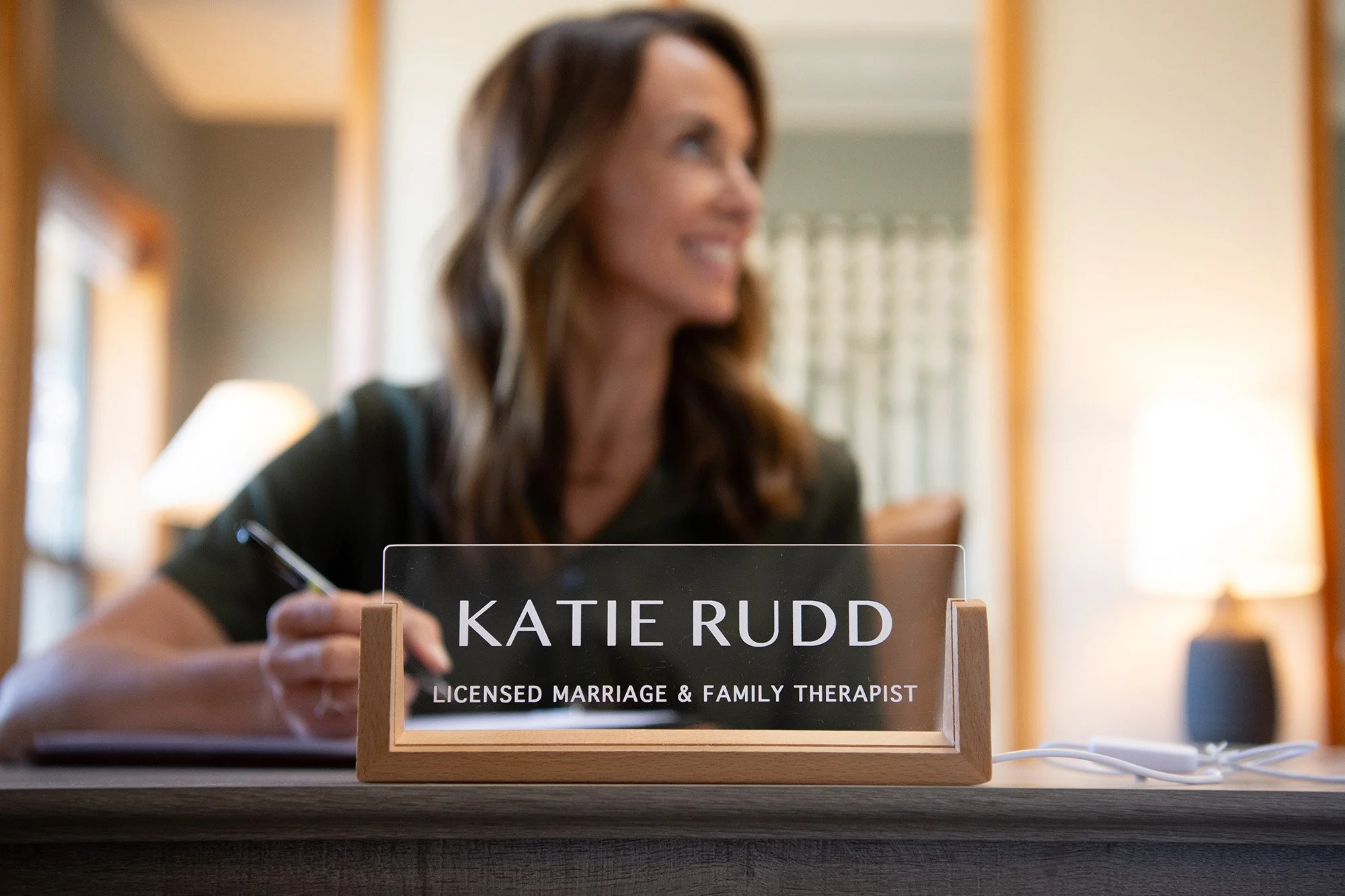 Close-up of a desk sign displaying the name "KATIE RUDD" and the title "Licensed Marriage & Family Therapist" in a professional office setting. Behind the sign, there is a smiling woman with long brown hair, slightly out of focus, writing on a notepa