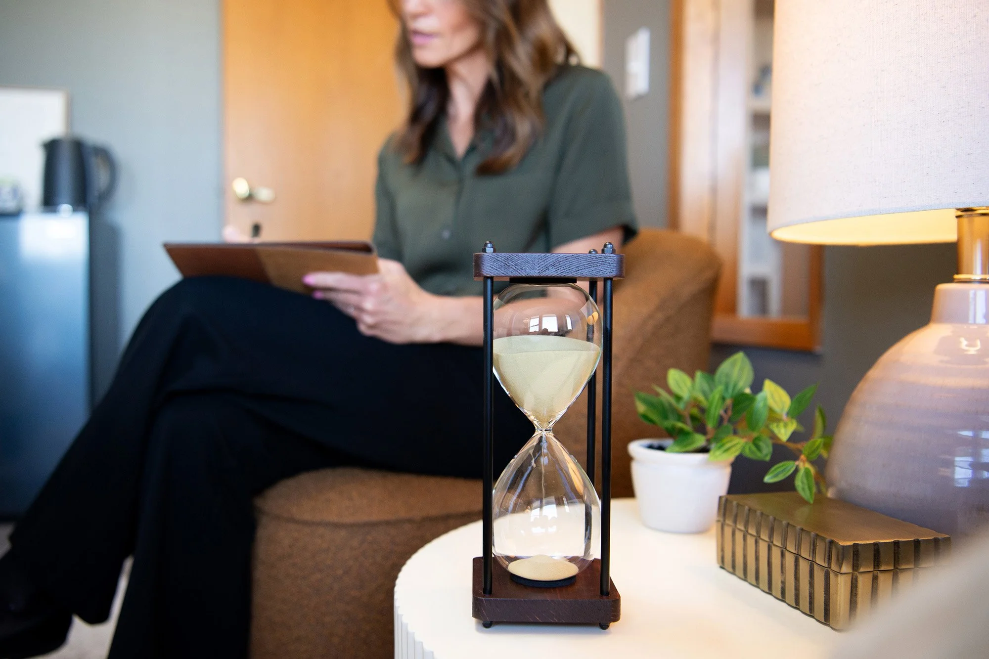 An hourglass on a white table in a cozy room with a woman sitting in the background, holding a tablet.