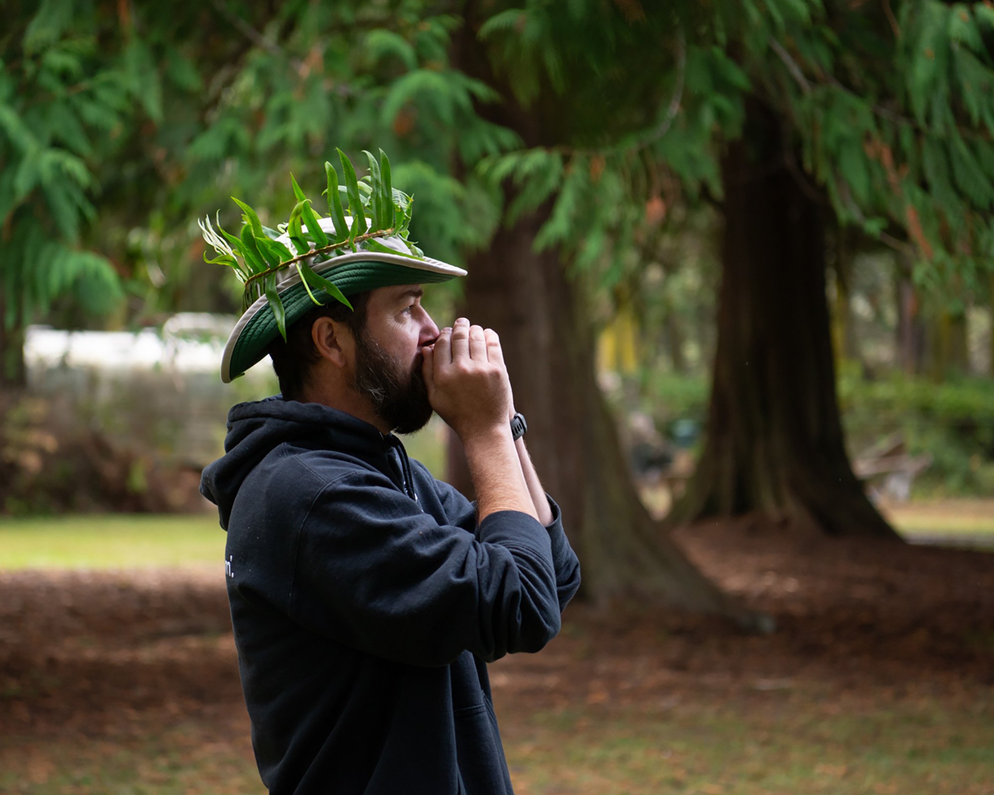 A man with a beard, wearing a black hoodie and a hat made of leaves, is standing outdoors in a wooded area, holding his hands near his face in a prayerful or meditative pose.