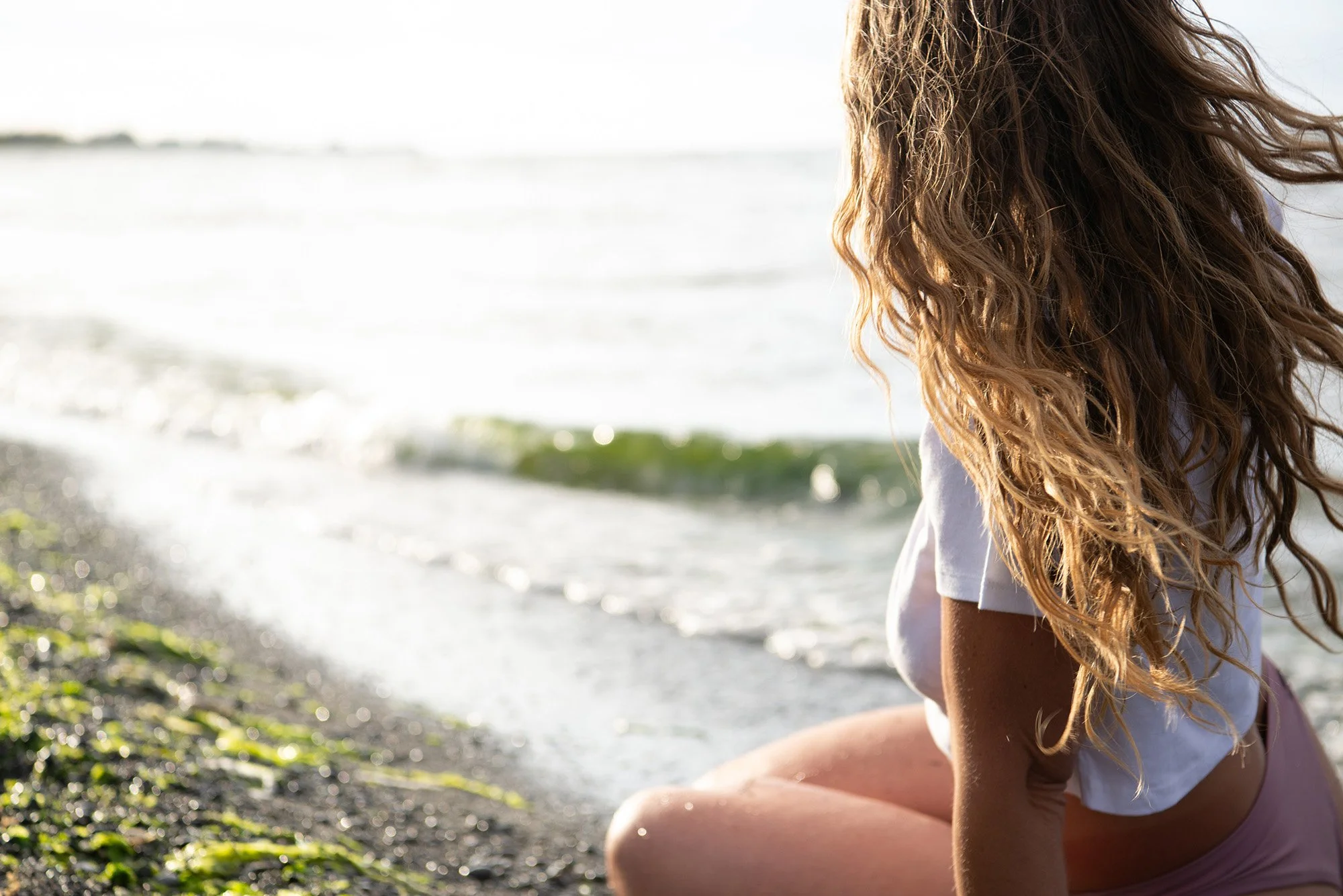 A woman with long curly hair sitting on a beach with green moss on rocks, facing the ocean during daytime.