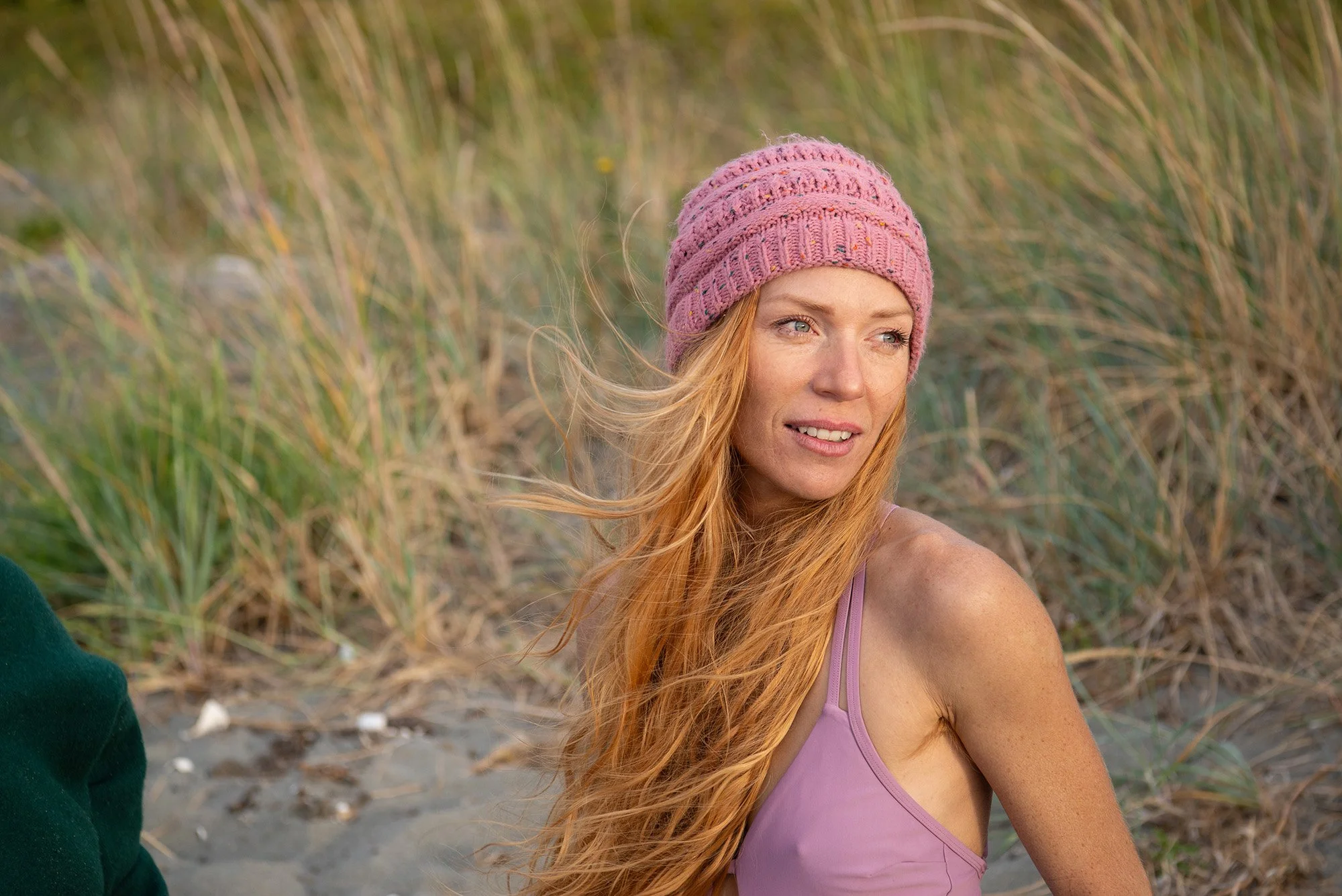 A woman with long red hair, wearing a pink knitted hat and purple tank top, sitting outdoors near grass and sand, looking to the side.