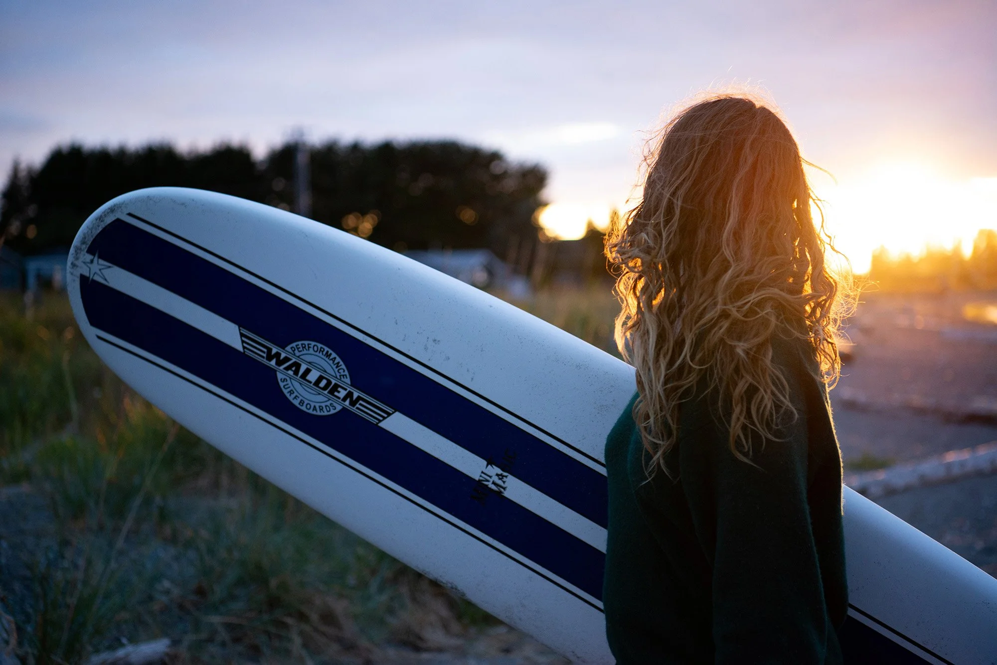 A woman with long curly hair holding a surfboard at sunset on the beach.