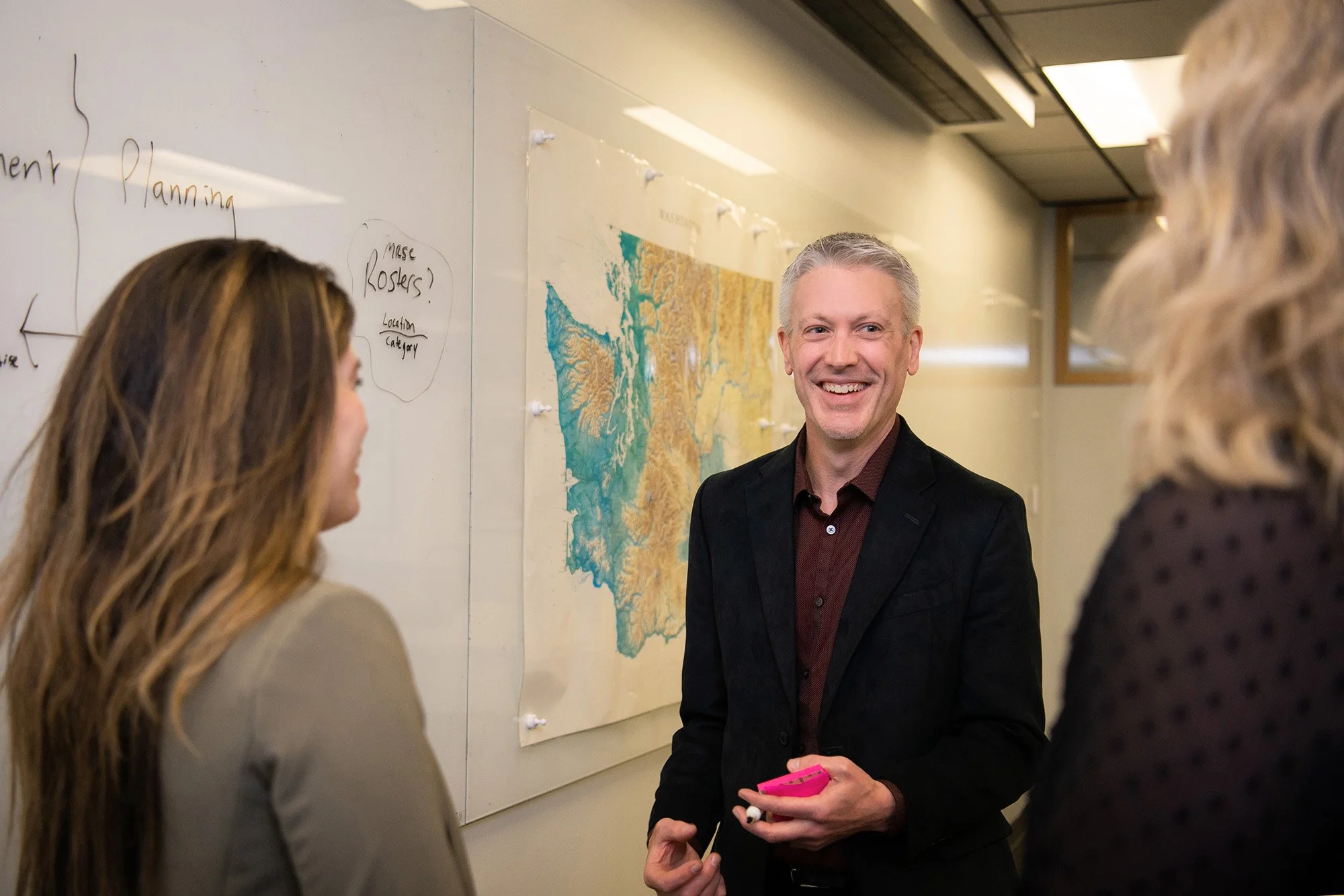 Three people talking in an office hallway, one man is smiling and holding a pink phone, a map and notes are on the wall.