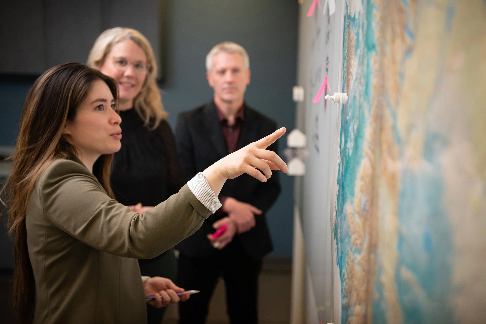 A woman in a beige blazer pointing at a large map on a wall, with two professionals observing.