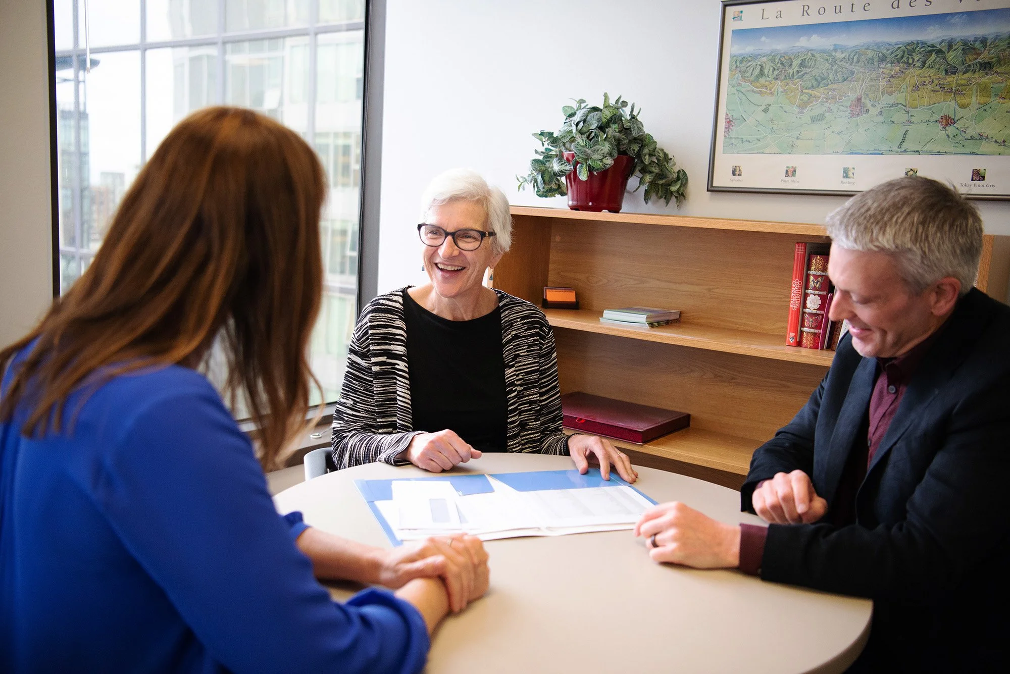 Three people sitting at a table in a meeting room, smiling and talking, with documents in front of them. A woman with gray hair and glasses is in the center, wearing a black and white striped cardigan, and two others, a woman with long brown hair in 