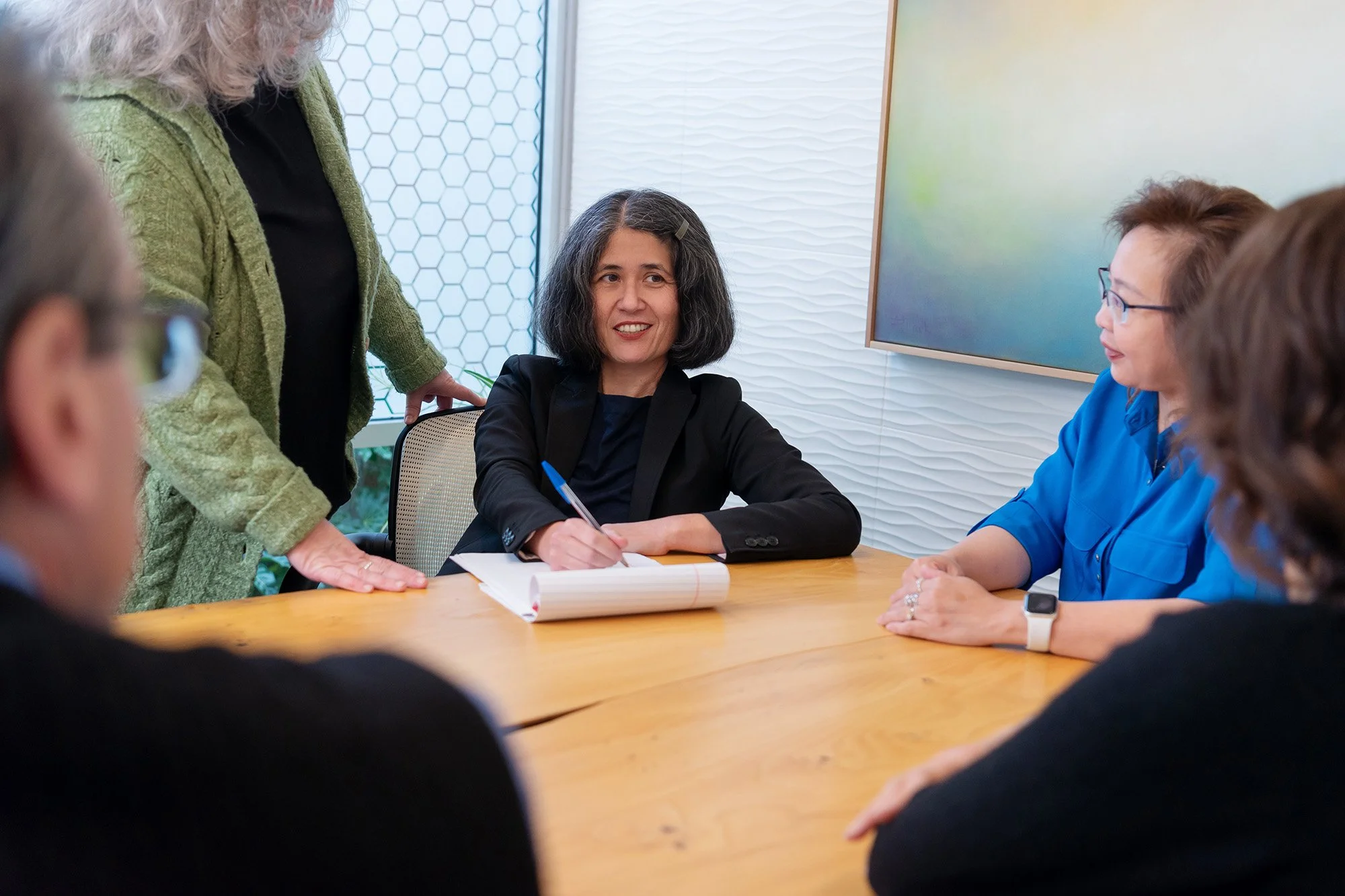 A woman with dark hair, wearing a black blazer, sitting at a conference table, writing in a notebook. Other women are sitting and standing around the table in a meeting room, with a colorful abstract artwork on the wall.