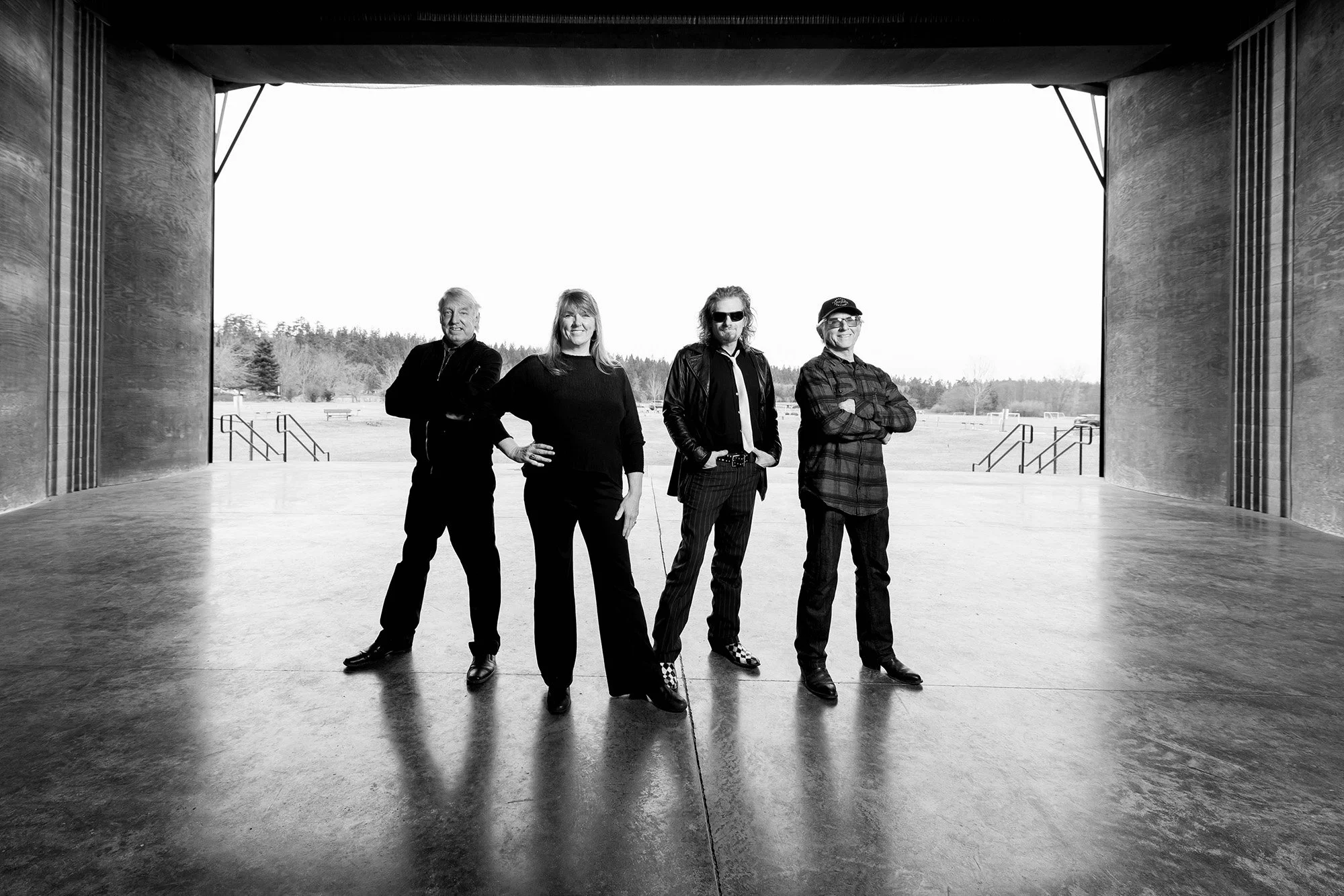 Four people standing under a large open structure outdoors with a grassy field and trees in the background, black and white photograph.