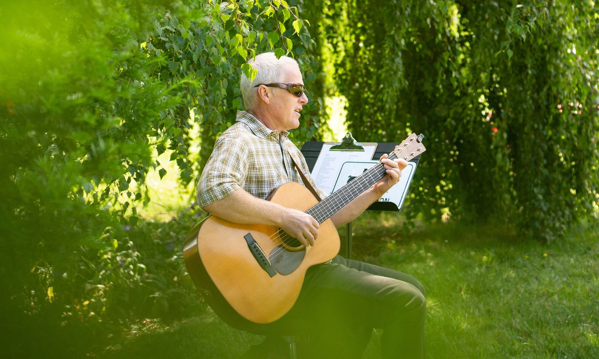An older man with white hair, wearing sunglasses and a plaid shirt, sitting outdoors and playing an acoustic guitar with a music stand next to him.