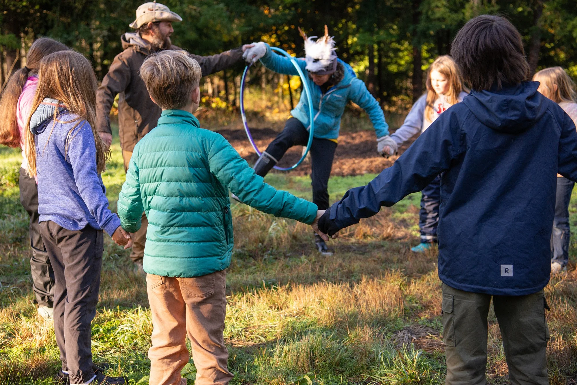 Children holding hands in a circle outdoors, with a woman in an animal costume wearing a unicorn horn and hula hoop in the background.