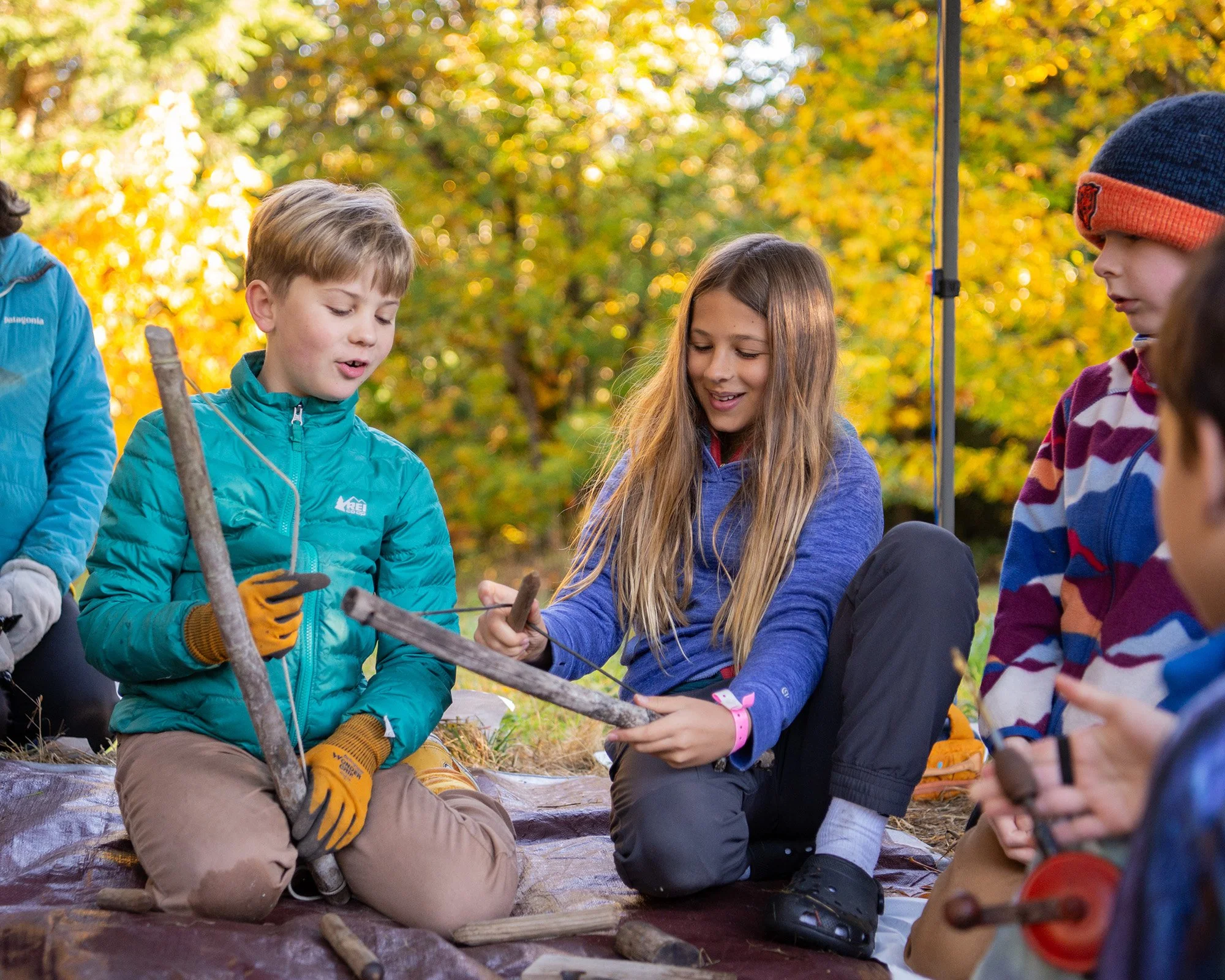 Group of children sitting outdoors in a forest during fall, participating in a fire-making activity with sticks and a fire starter, with autumn foliage in the background.