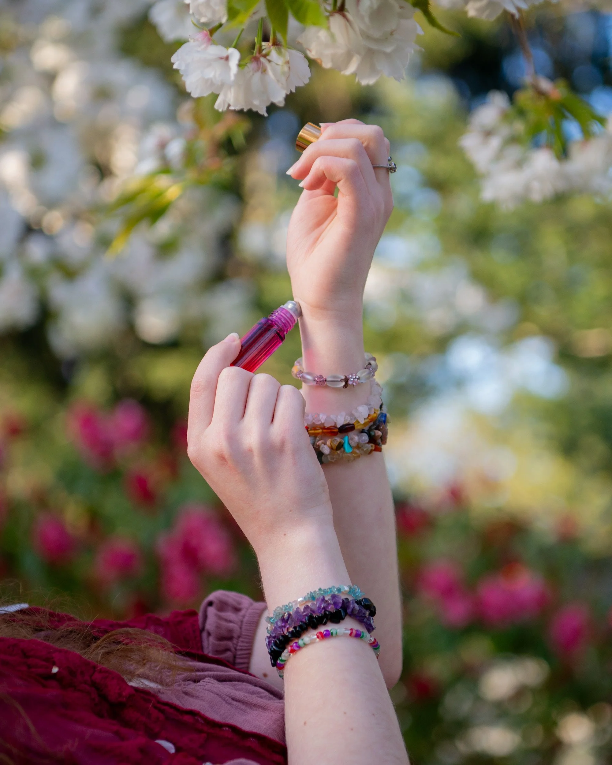 A person holding a perfume bottle near blooming cherry blossoms outdoors, wearing multiple beaded bracelets on their wrist.