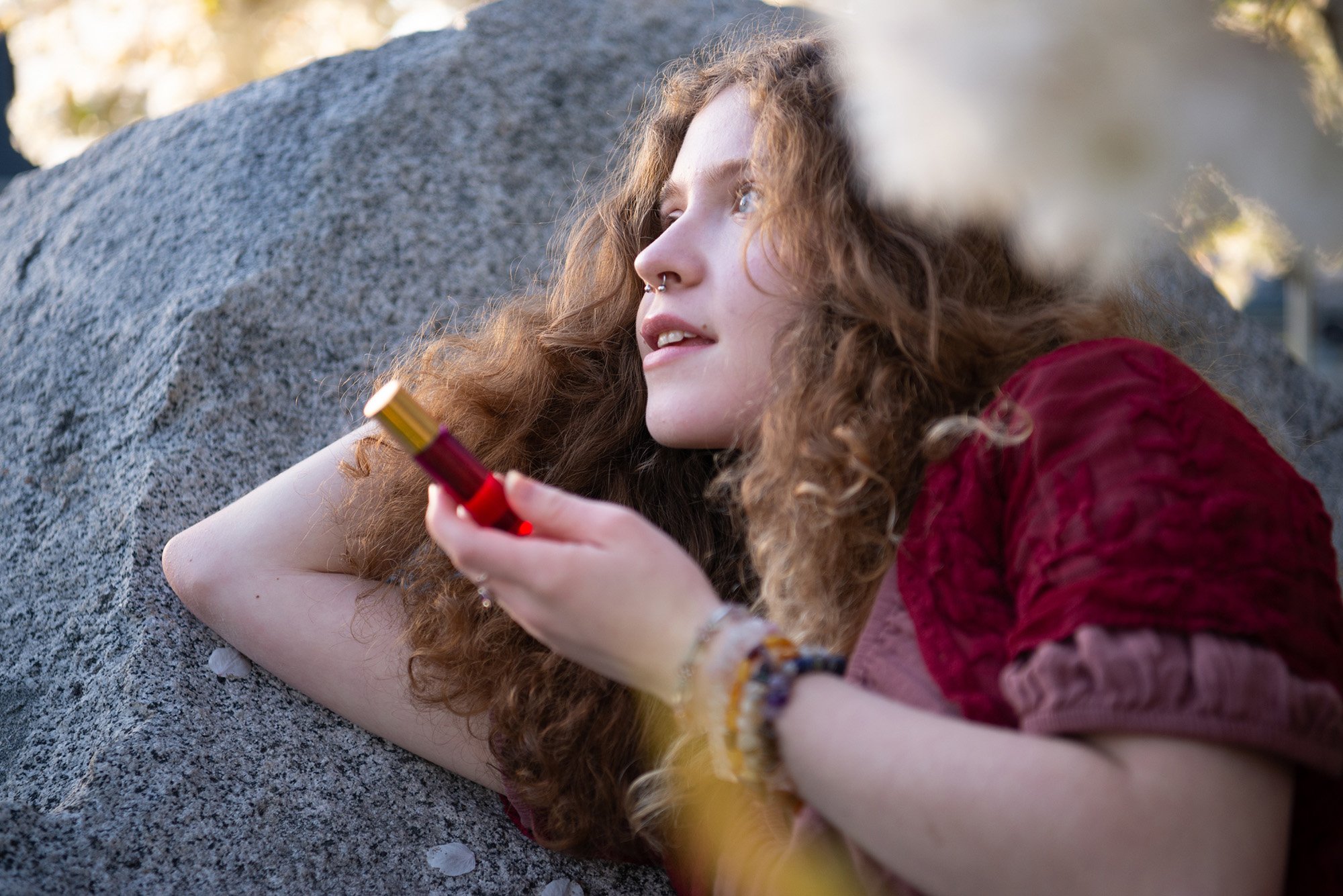 Young woman with curly hair lying on a rock holding a small bottle, outdoors with sunlight