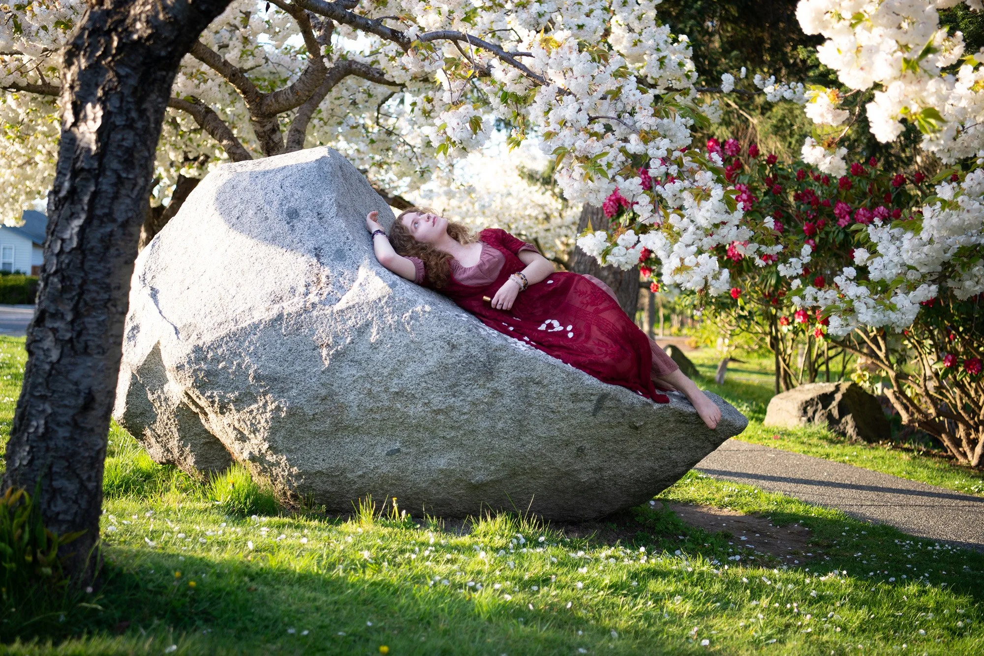 A woman in a red dress lying on a large gray rock surrounded by blooming white and pink flowers on trees.
