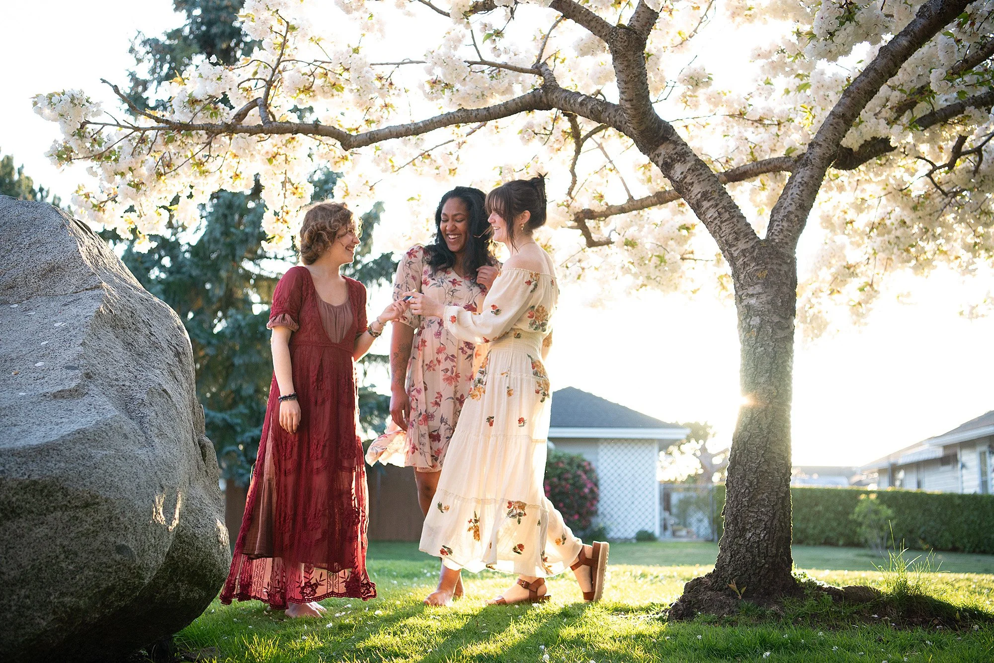 Three women enjoying spring outdoors under flowering tree, smiling and chatting, with house and greenery in background.