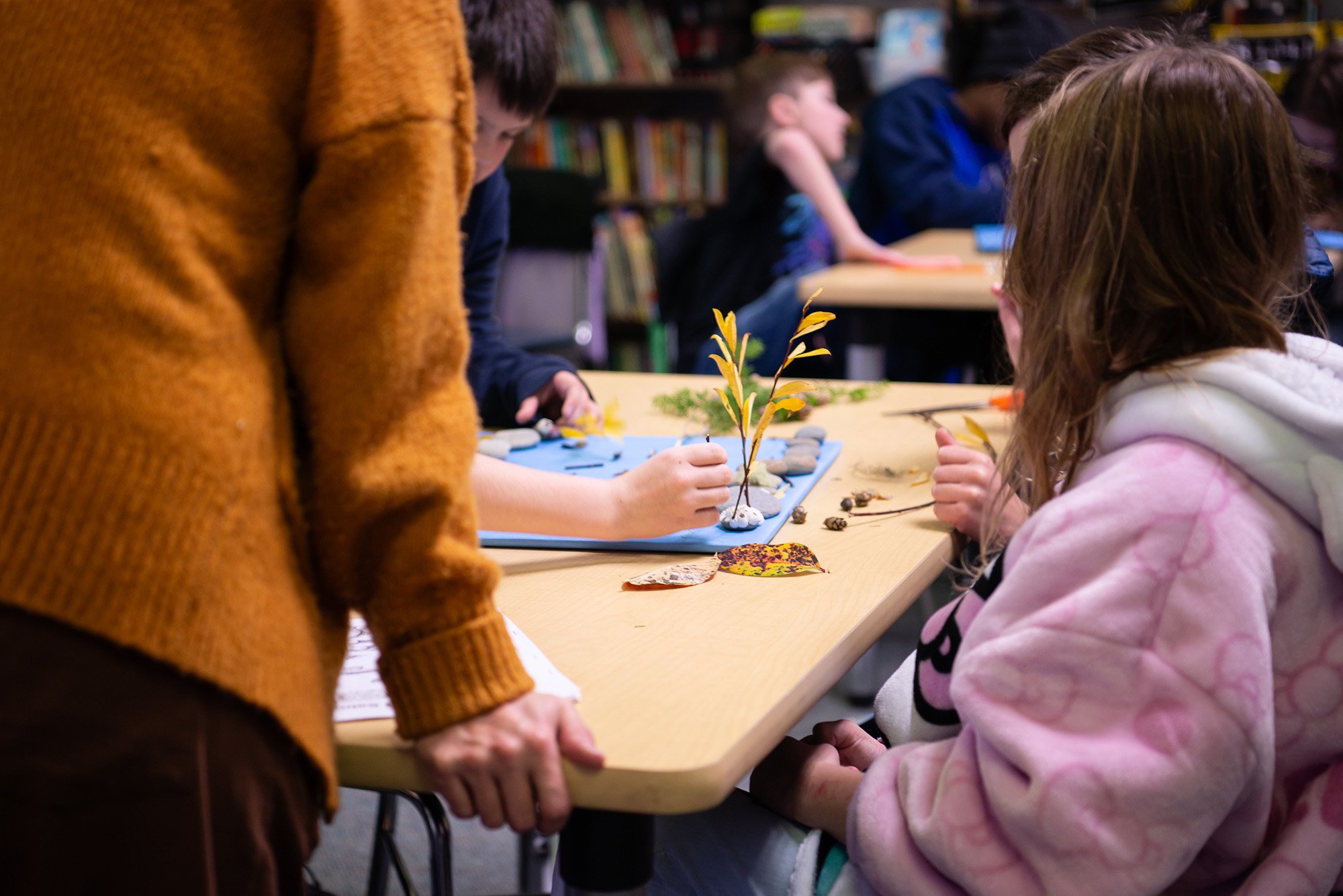 Children and an adult working on a nature-based craft project with leaves, rocks, and small plants at a table in a classroom or library.