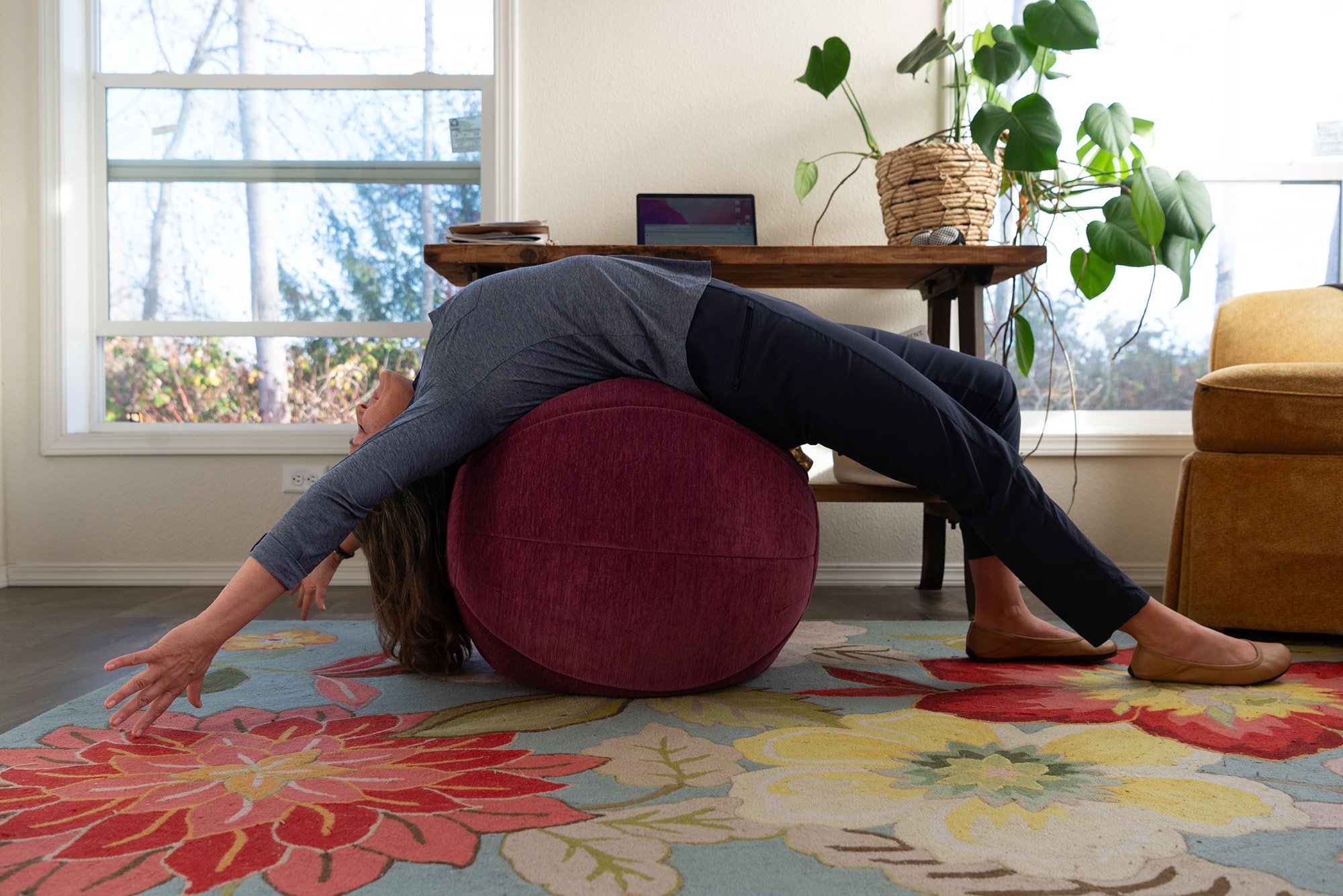 A woman stretching over a round, purple cushion in a living room with large windows, a wooden desk with a tablet, a potted plant, and a yellow armchair.