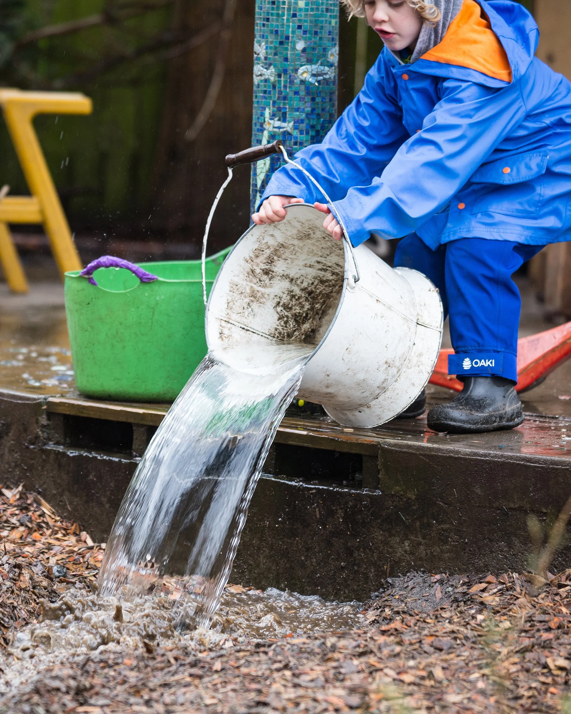 Child in blue raincoat pouring water from a bucket into a small stream in a backyard.