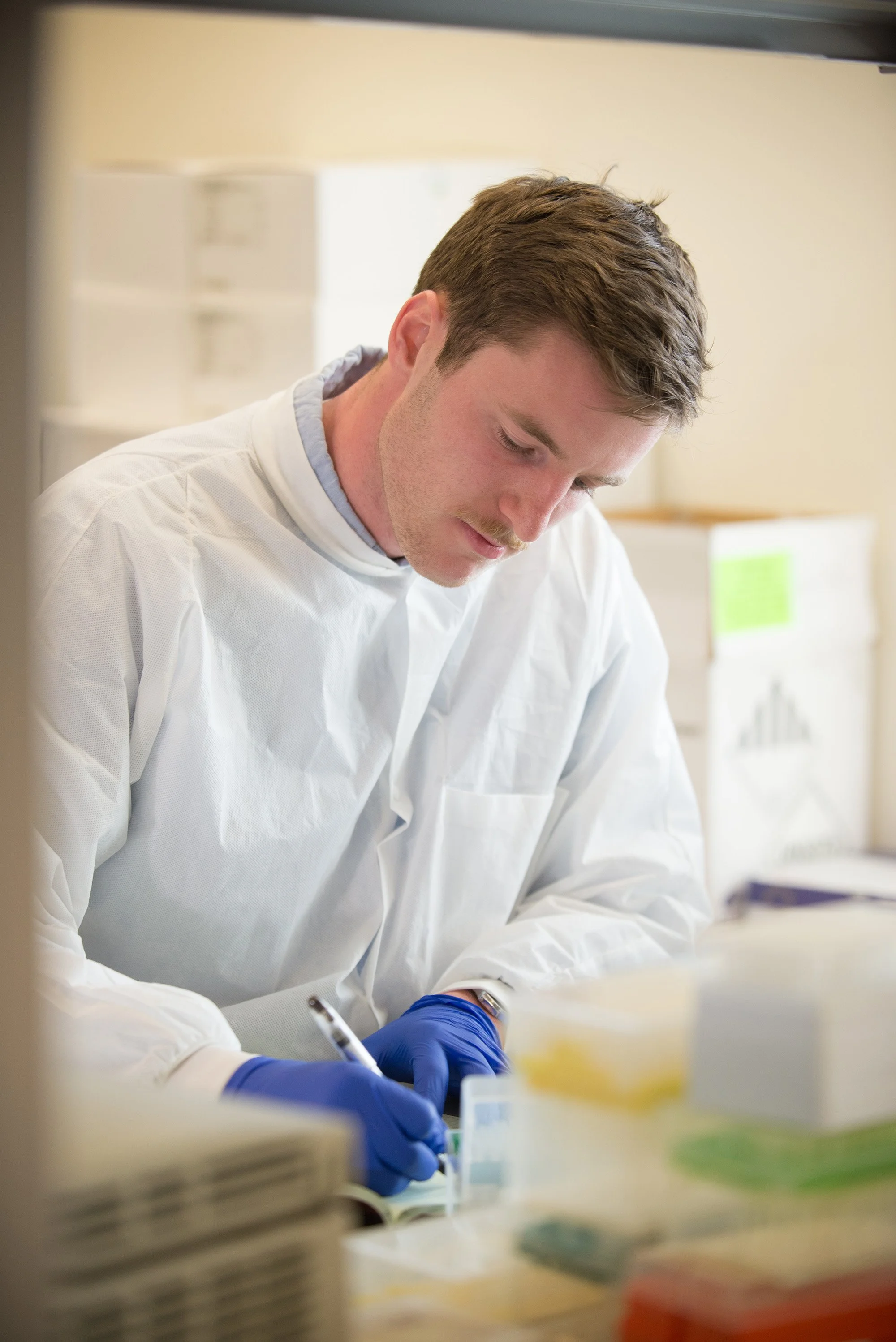 A scientist wearing a white lab coat and blue gloves is working in a laboratory, writing notes on a clipboard.