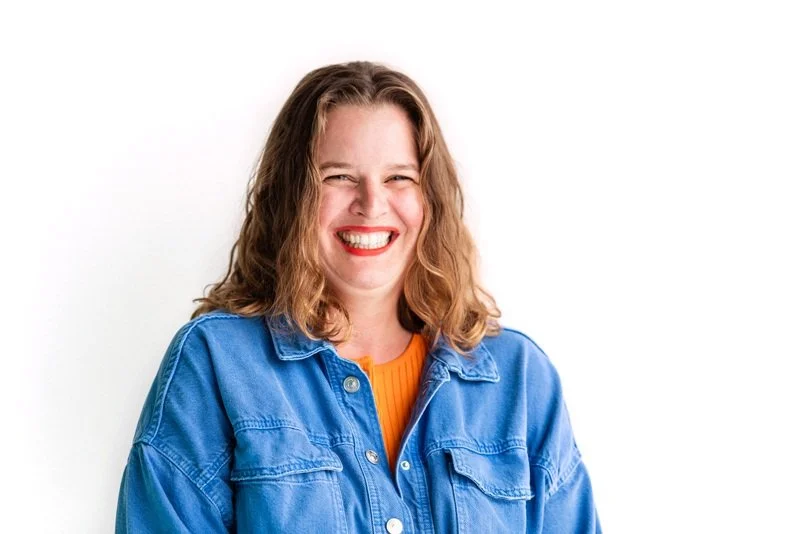 Portrait of Jennifer Hennesy, artist, art director and graphic designer. Jennifer is a white woman with a huge smile on her face. Wavy shoulder-length brown hair, red lipstick, a blue denim jacket with a bright orange shirt