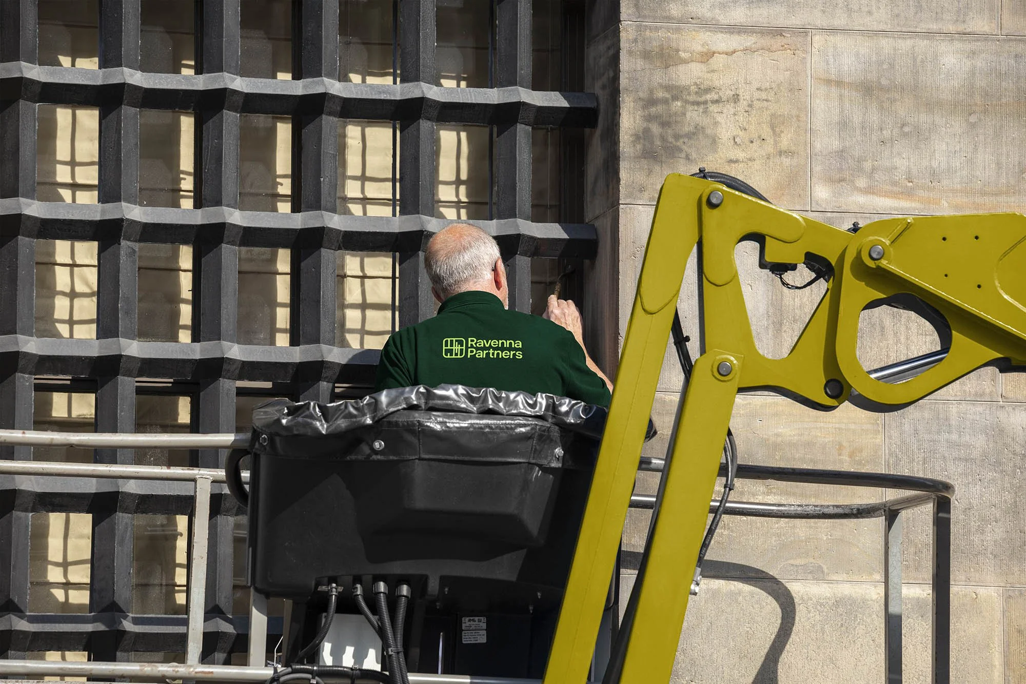 A man with gray hair is working on a large, black metal window grille on the exterior wall of a building. He is wearing a green jacket with the logo and words 'Ravenna Partners' on the back. The man is on a yellow construction lift platform, which is partially visible in the foreground.