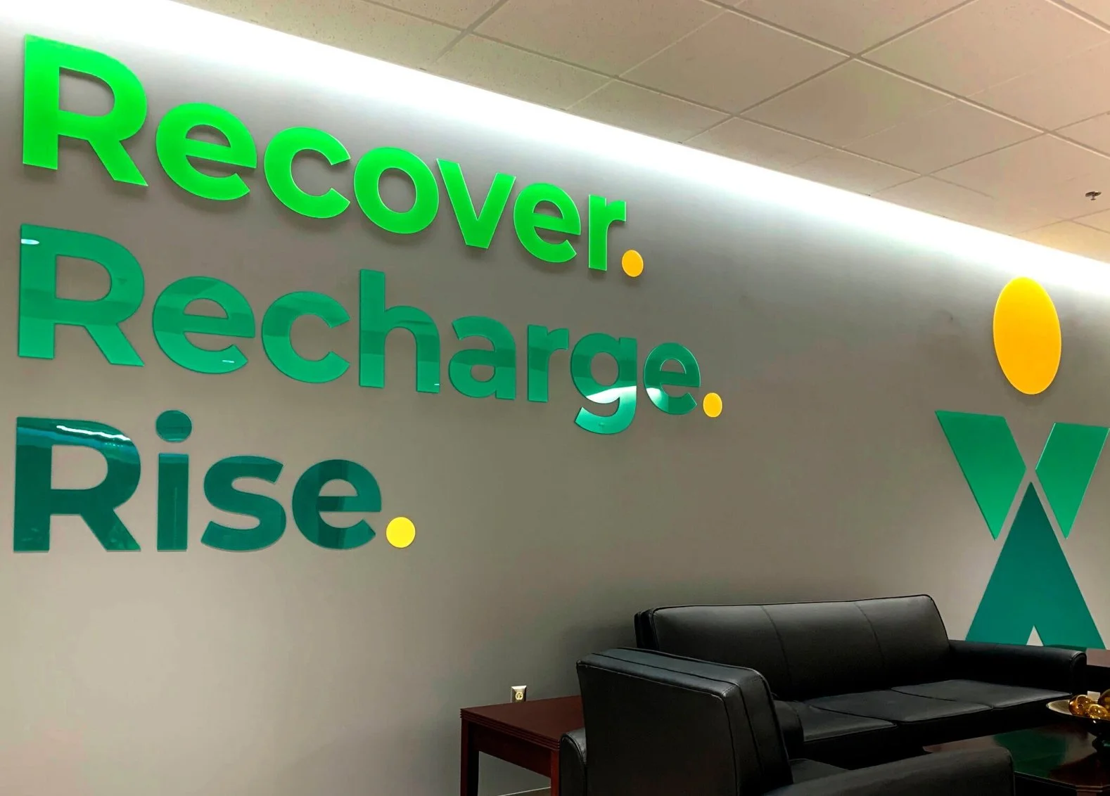 An office lobby wall with motivational words in large green letters: "Recover. Recharge. Rise." A stylized logo with a yellow circle and a blue and green abstract X is on the right. There is a black leather sofa, a side table, and a decorative bowl with gold balls.