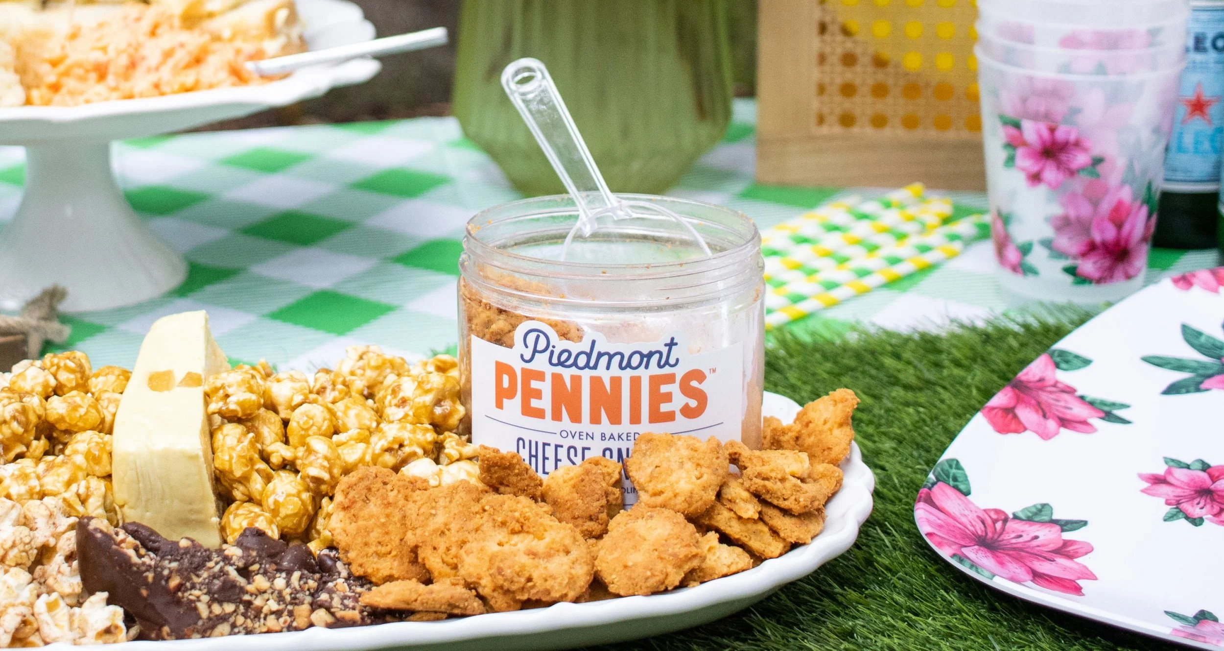 A plate of caramel popcorn, cheese popcorn, chocolate-covered popcorn, and fried chicken nuggets on a picnic table with a jar of Piedmont Pennies cheese balls in the center.
