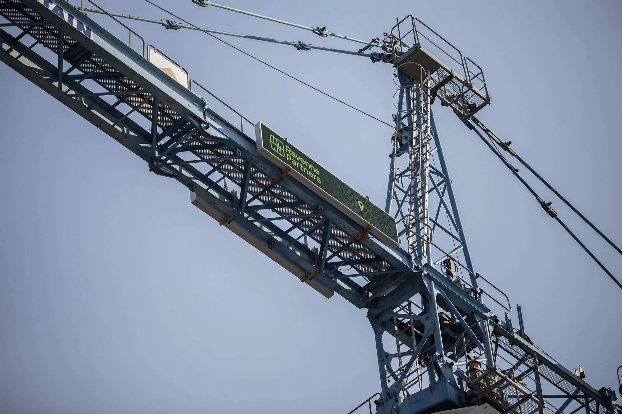 Close-up of a blue construction crane against a clear sky, with a digital sign displaying 'Ravenna Partners'.