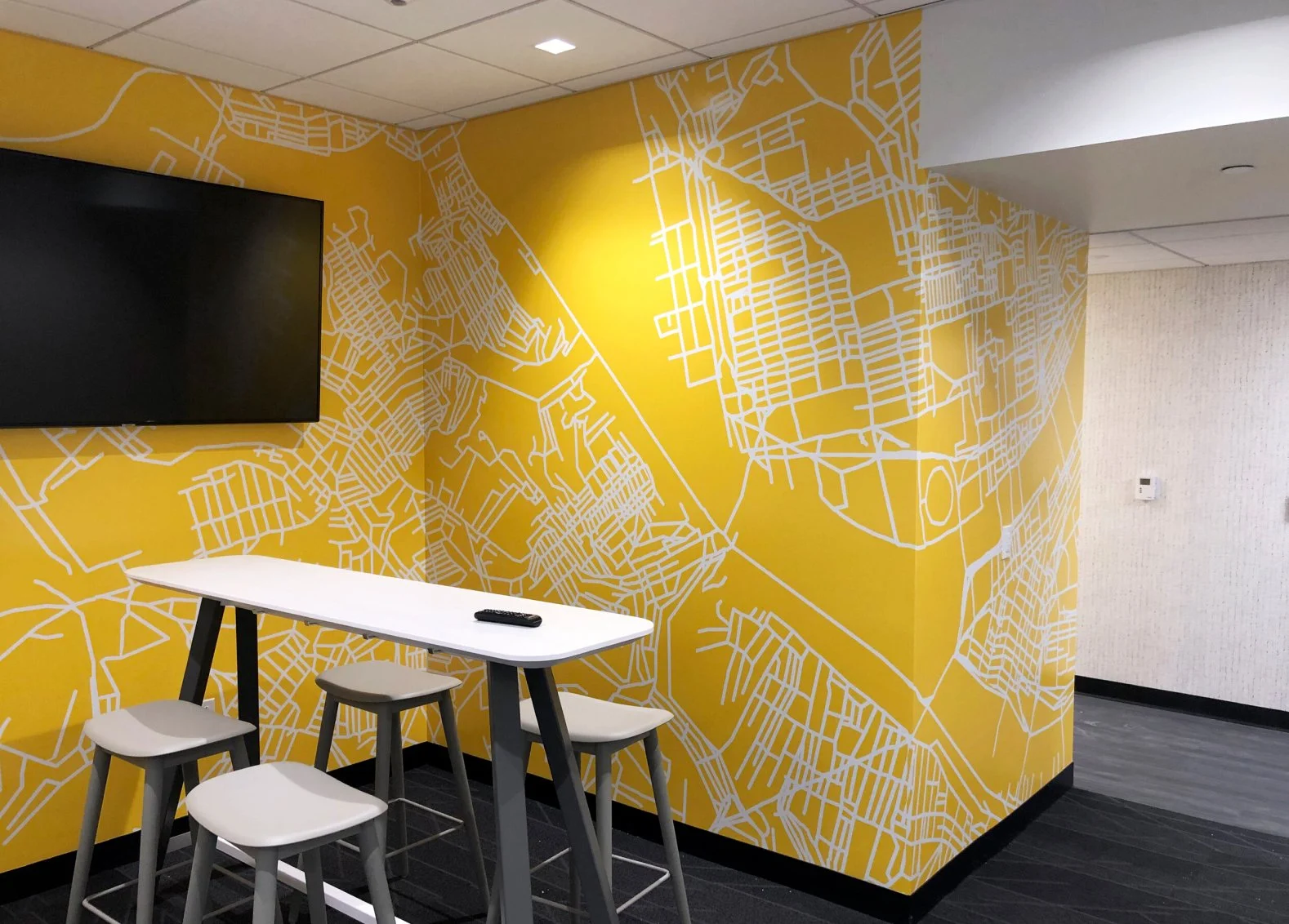 Yellow wall with white map design, a large black television, a high table with three white stools, and a remote control on the table. The space has a modern look.