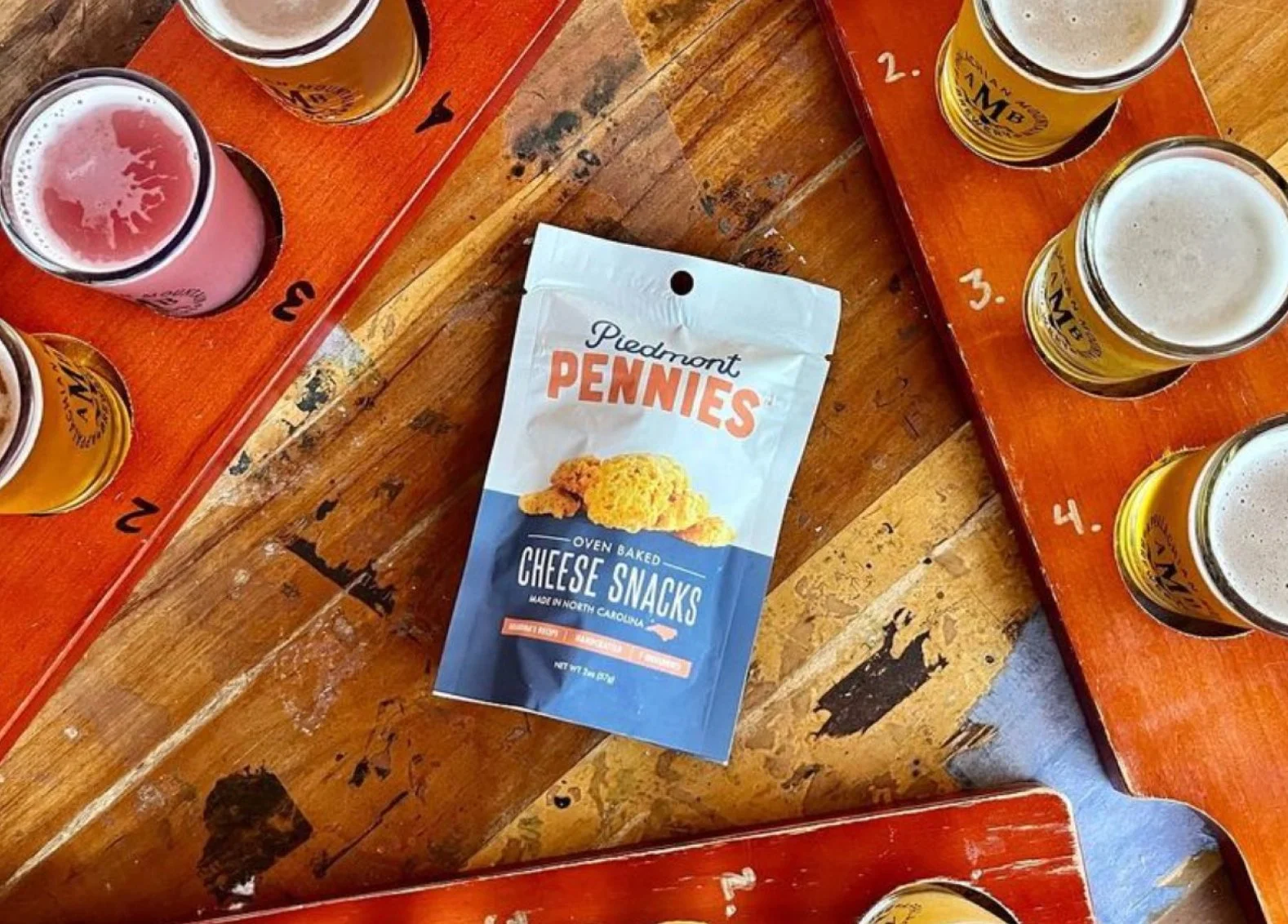 A wooden table with a bag of Piedmont Pennies cheese snacks in the center and four flights of beer tastings on red trays surrounding the bag.