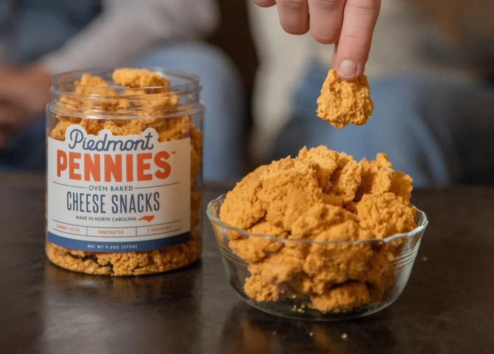 A person is grabbing a cheese cracker from a bowl of cheese crackers labeled 'Piedmont Pennies Cheese Snacks'. The crackers are in a glass jar on a table, with the jar's label visible.