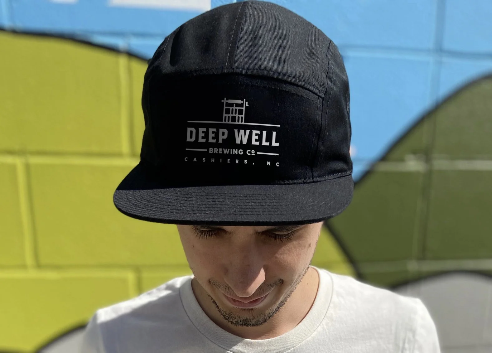 Person wearing a black cap with 'Deep Well Brewing Co' logo, standing outdoors with colorful background.