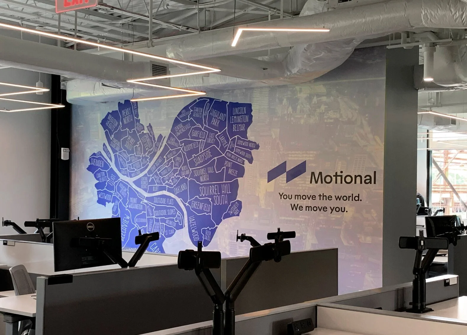 Office workspace with a wall map of a city in shades of blue, next to a slogan that says, 'You move the world. We move you.' The workspace has desks with monitors and adjustable arms, modern lighting, and an industrial ceiling.