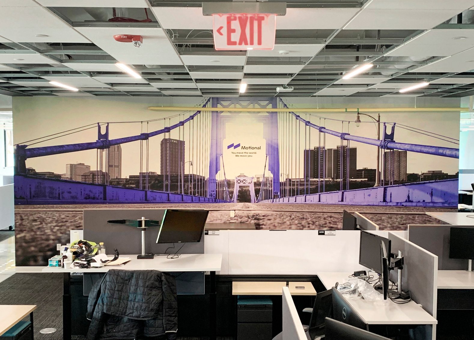 Office space with work desks and monitors, some clutter, and a wall mural of a suspension bridge with a city skyline in the background, featuring a central sign with 'Motional' and the tagline 'You move the world. We move you.'