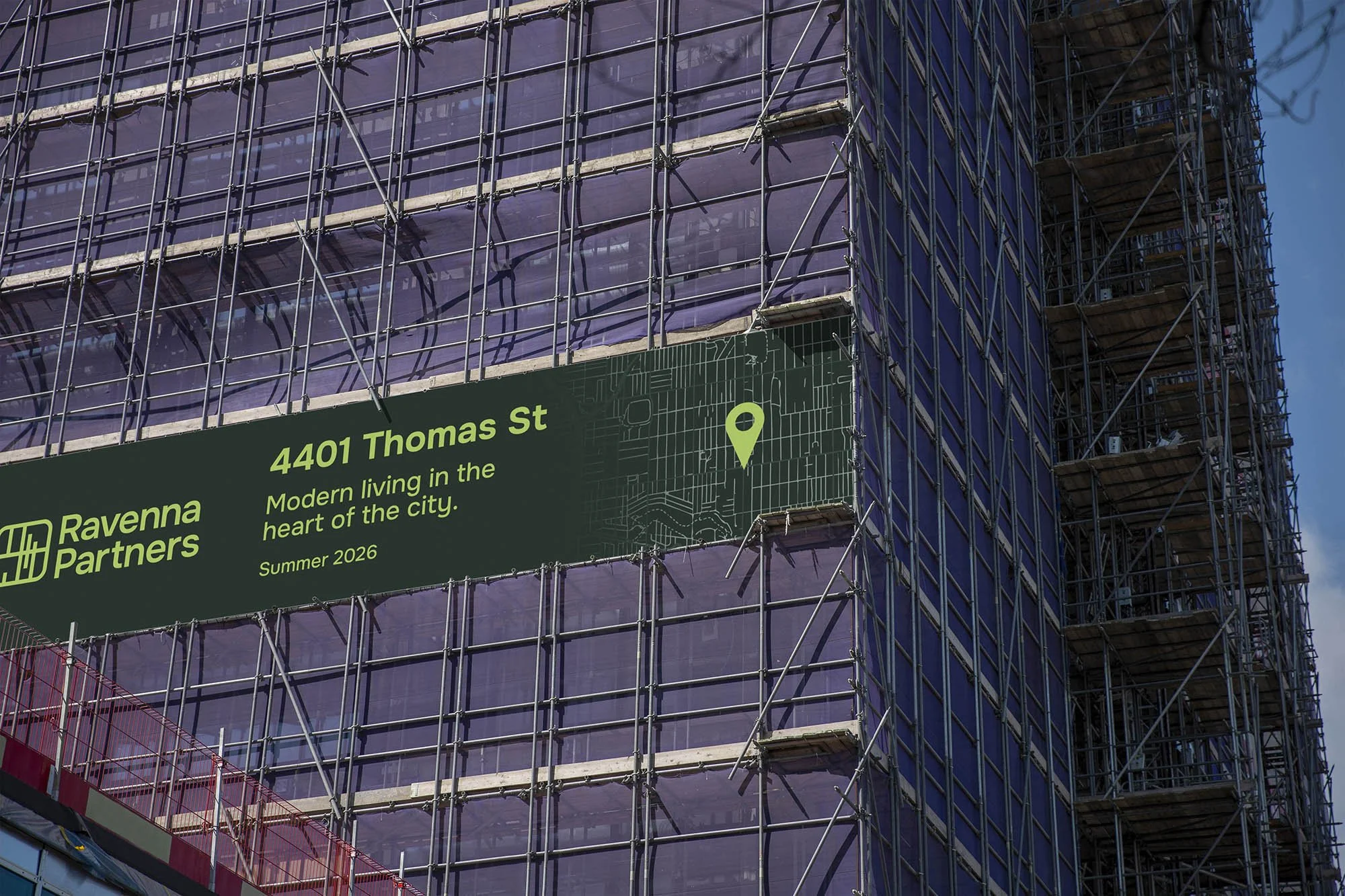 Construction site of a modern building with purple-tinted glass windows and scaffolding. A green sign on the building displays an address, '4401 Thomas St,' and promotional text about the development by Ravenna Partners, indicating it is 'Modern living in the heart of the city,' scheduled for summer 2026.