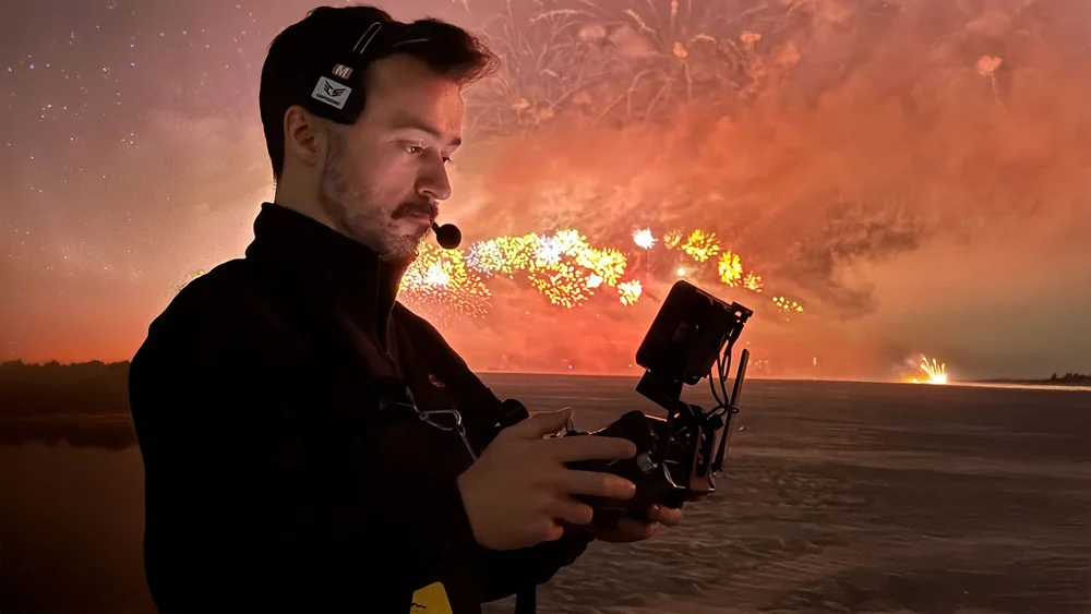 Drone pilot with a camera controller and headset watching a fireworks display over the water at night