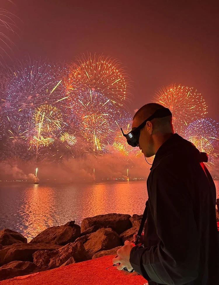 FPV pilot in goggles on the rocks by the water during a fireworks display