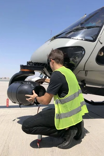 Technician rigging a Shotover F1 camera system under a helicopter on the apron