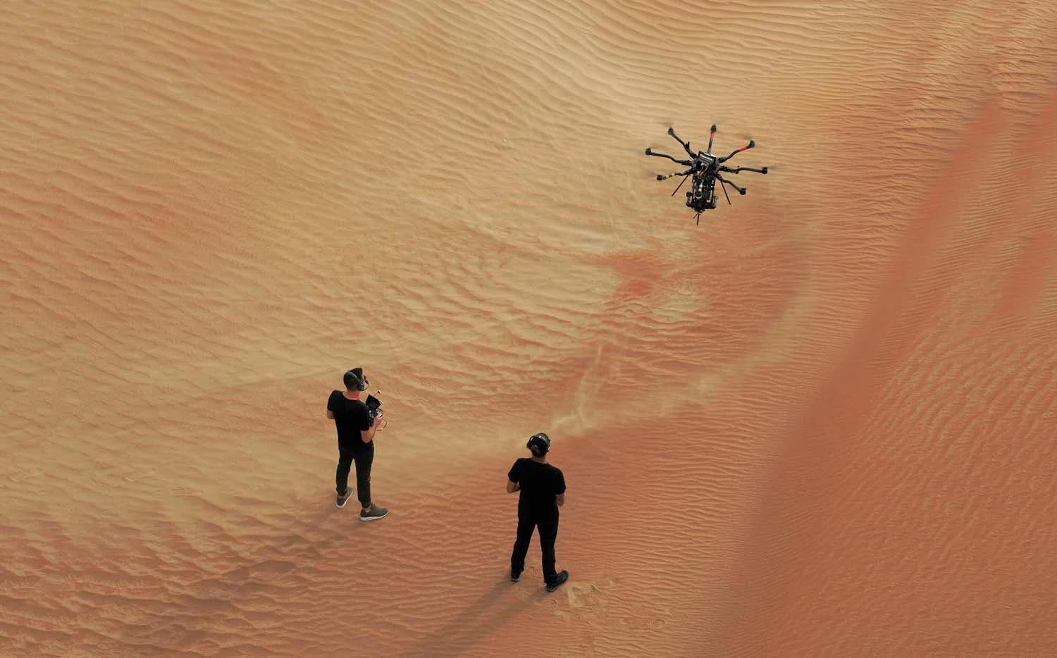 Top-down view of a heavy-lift cinema drone flying over desert sand, with the pilot and camera operator below