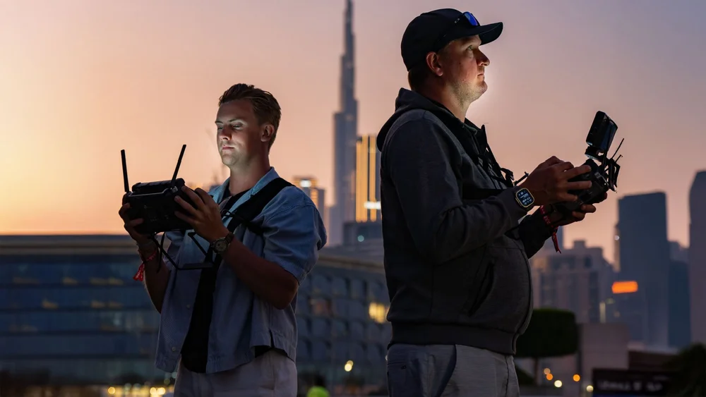 Two drone pilots holding remote controllers at dusk in Downtown Dubai with the Burj Khalifa behind them