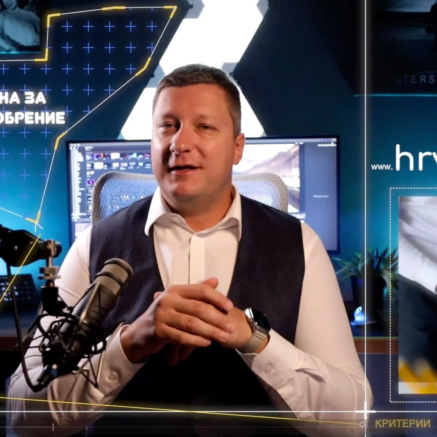 A man in a white shirt and black vest sitting in front of a computer screen, speaking into a microphone, with colorful graphics and text in a studio or conference setting.