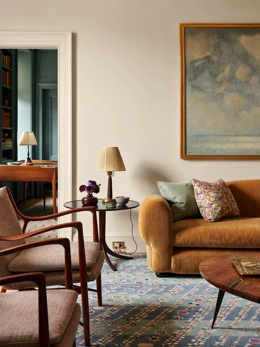 Living room with a mustard sofa, pink and green pillows, a wooden coffee table, and a side table with a lamp, decorative vase with purple flowers, and a bowl. There is a framed abstract painting on the wall and a doorway leading to a room with a wooden desk, chairs, and another lamp.