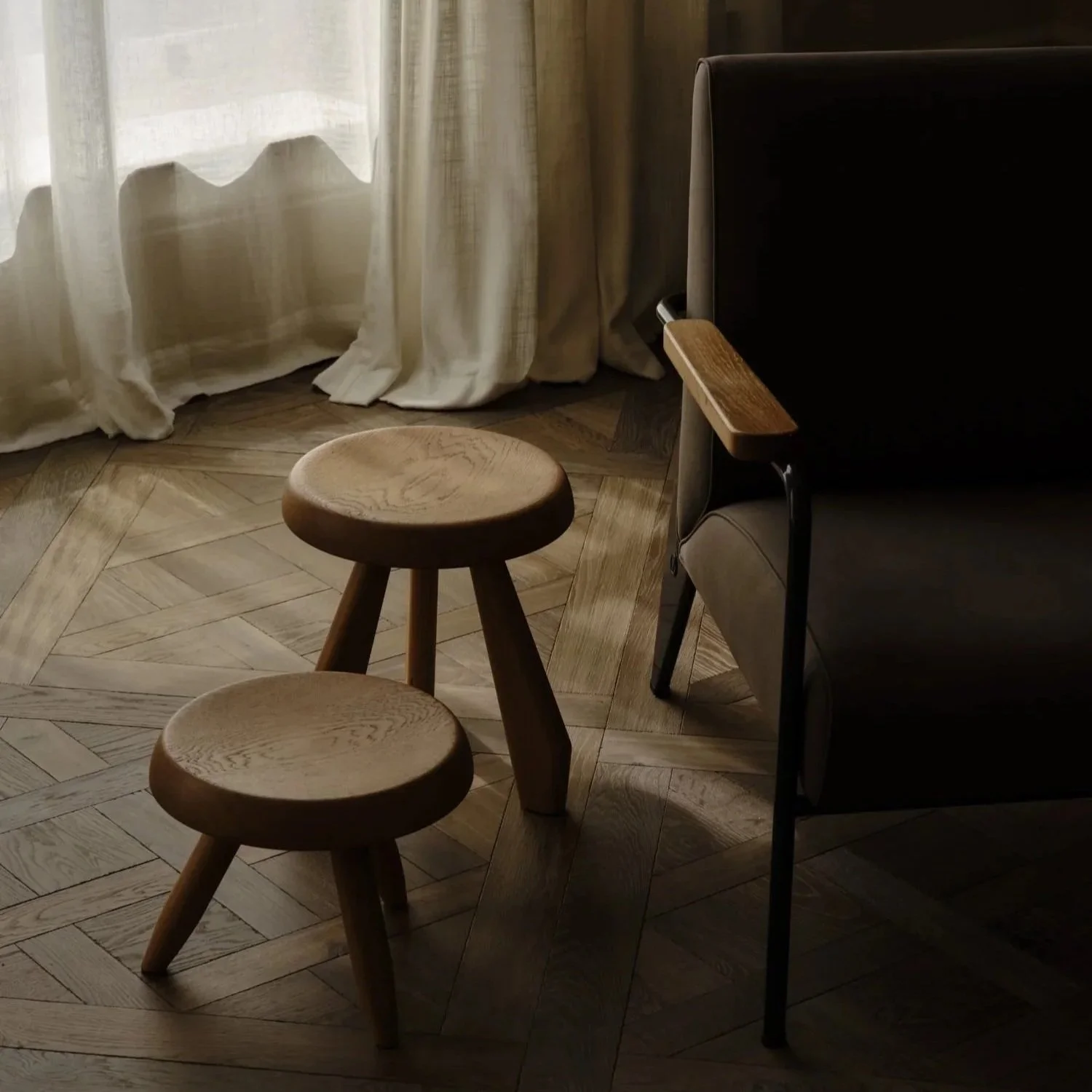 A corner of a room with wooden stools, a black armchair with a wooden armrest, light-colored curtains, and a wooden parquet floor.