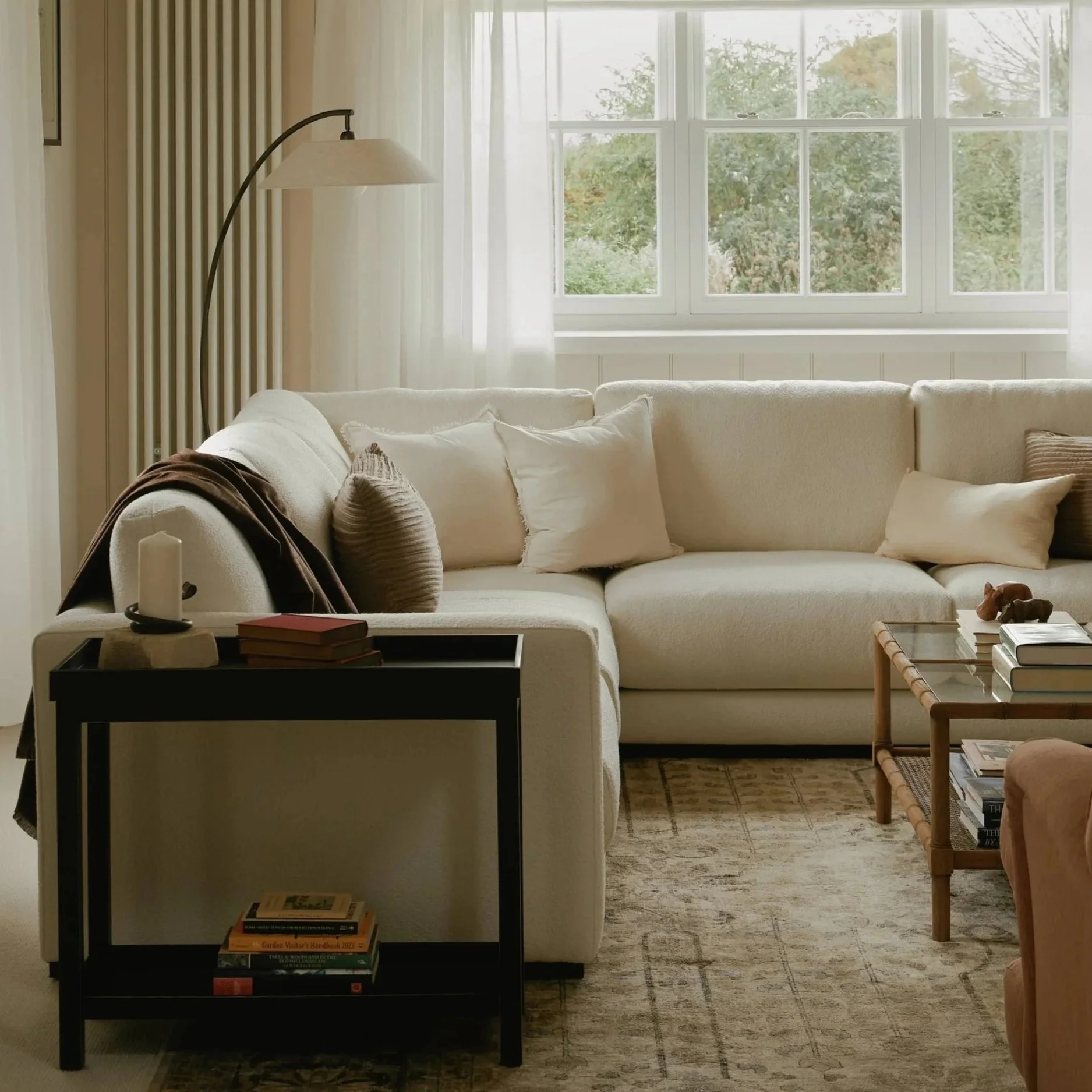 A cozy living room with a large window letting in natural light, a white sofa with pillows, a bookshelf with books, a side table with a candle, a small table with books and decorative items, and a patterned rug.