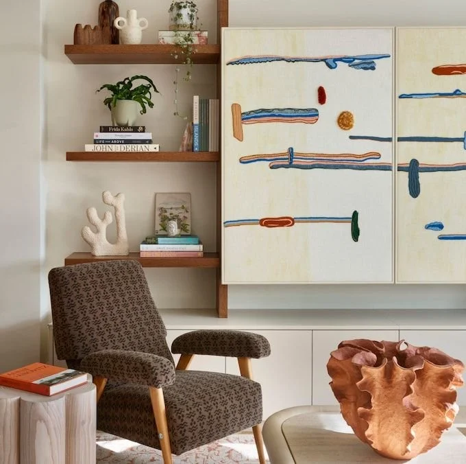 Interior lounge with a vintage armchair and media wall to rear including wooden shelves with books and decorative items. A large abstract art piece conceals the television, with a textured clay vase on a table.