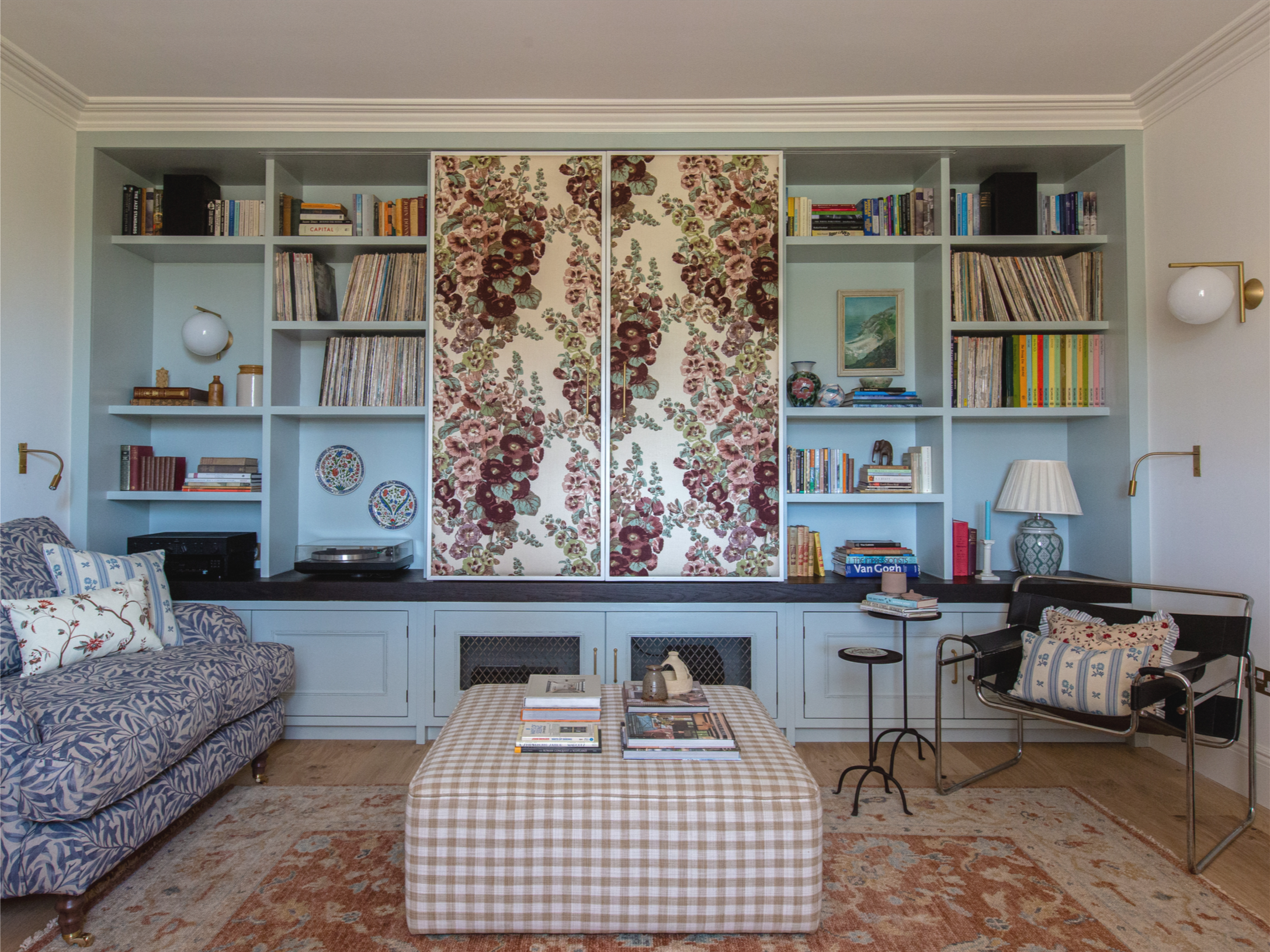 A living room with a large bookshelf, floral sliding doors, a blue patterned sofa, a plaid ottoman, side tables, lamps, books, and decorative items.