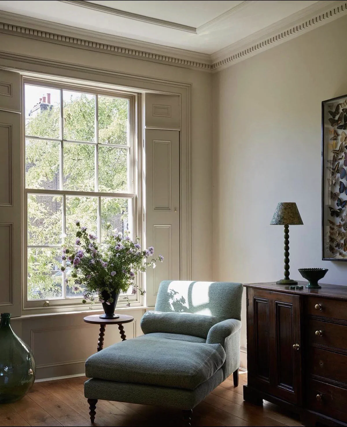A cozy living room corner with a large window, a light green armchair with an ottoman, a wooden sideboard with a lamp and decorative bowl, a framed butterfly art on the wall, and a vase of flowers on a small table.